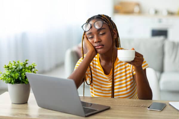 Can Caffeine Cause Insomnia? Exhausted woman sitting in front of a laptop with a cup of coffee in her hand