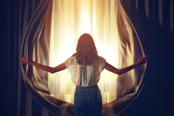 Back view of a woman opening curtains at home with the sun shining through