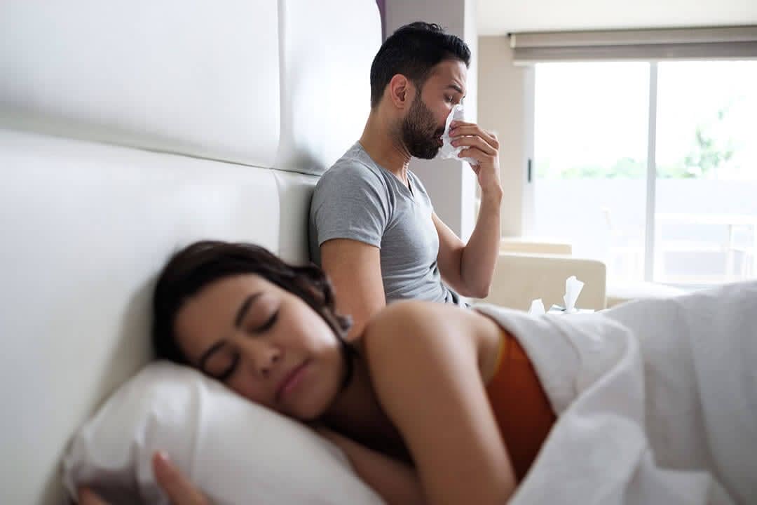 How to Sleep with Allergies Young Man with Allergies Blows Nose in Bed While Female Partner Sleeps